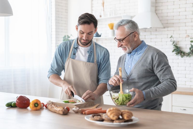 Un jeune homme et un monsieur aux cheveux gris préparent une salade ensemble, en souriant