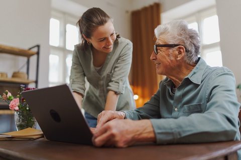 Une jeune femme aide un monsieur âgé à effectuer une démarche sur un ordinateur portable, à son domicile