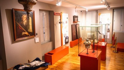 Salle d'exposition du musée : vitrines orange présentant des couronnes de Torah en argent, tableau ancien d'un patriarche tenant les Tables de la Loi