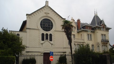 Façade claire de la synagogue de Biarritz, avec sa grande rosace, entre palmier et villa à tourelle
