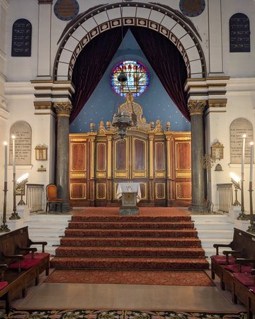 Intérieur de la synagogue de Bayonne : l'arche sainte en bois sous une voûte bleue et un vitrail, au sommet de marches recouvertes d'un tapis rouge